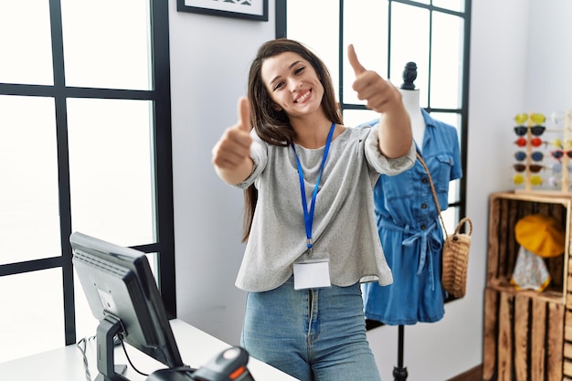 Young brunette woman working as manager at retail boutique approving doing positive gesture with hand thumbs up smiling and happy for success winner gesture