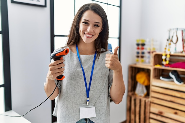 Young brunette woman working at retail store holding barcode reader device smiling happy and positive, thumb up doing excellent and approval sign