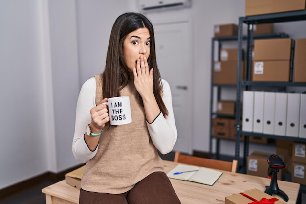 Young brunette woman working at small business ecommerce holding i am the boss cup covering mouth with hand, shocked and afraid for mistake. surprised expression