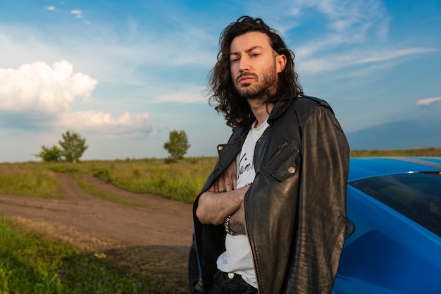 A young brutal man with long hair and bristles stands near a bright blue car on a middle of a rural road.