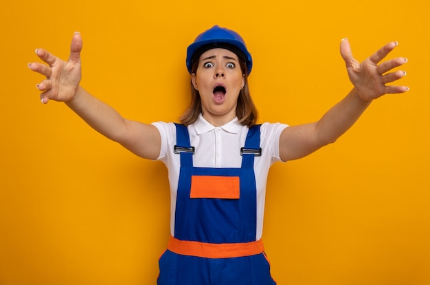 Giovane donna costruttore in uniforme da costruzione e casco di sicurezza scioccata alzando le braccia in preda al panico in piedi sul muro arancione