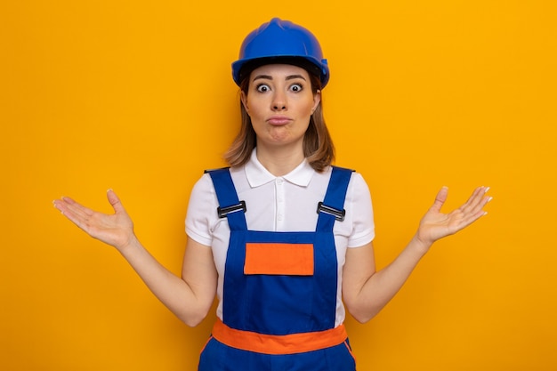 Young builder woman in construction uniform and safety helmet confused spreading arms to the sides having no answer standing on orange