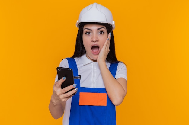 young builder woman in construction uniform and safety helmet holding smartphone