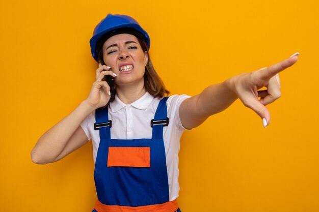 Young builder woman in construction uniform and safety helmet looking aside annoyed and irritated pointing with index finger at something while talking on mobile phone standing over orange wall