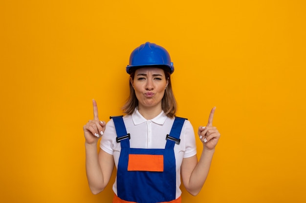 Young builder woman in construction uniform and safety helmet looking confused showing large size gesture with hands and fingers