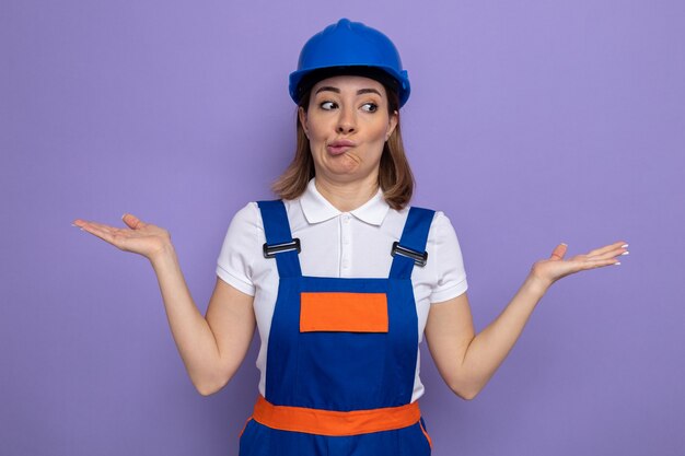 Young builder woman in construction uniform and safety helmet looking confused shrugging shoulders standing on purple