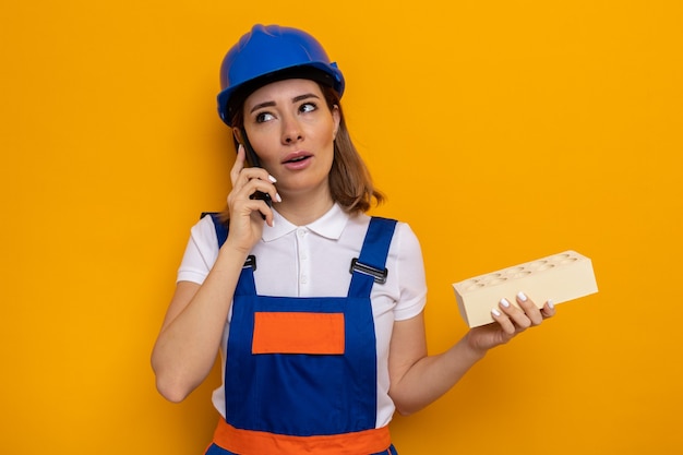 Young builder woman in construction uniform and safety helmet looking confused while talking on mobile phone holding brick standing over orange wall