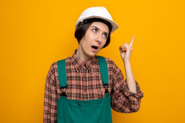 Giovane donna del costruttore in uniforme da costruzione e casco di sicurezza che guarda in alto perplesso mostrando il dito indice in piedi sull'arancia