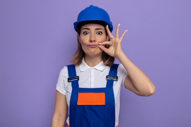 건설 유니폼을 입은 젊은 건축업자 여성과 보라색 벽 위에 지퍼가 서 있는 입을 닫는 것과 같은 침묵의 몸짓을 하는 안전 헬멧