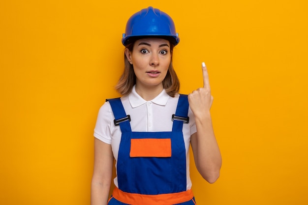 Young builder woman in construction uniform and safety helmet  surprised showing number one index finger standing over orange wall