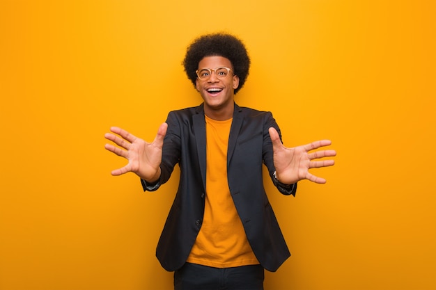 Young business african american man over an orange wall very happy giving a hug to the front