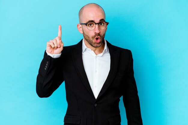 Young business bald man isolated on blue wall pointing upside with opened mouth