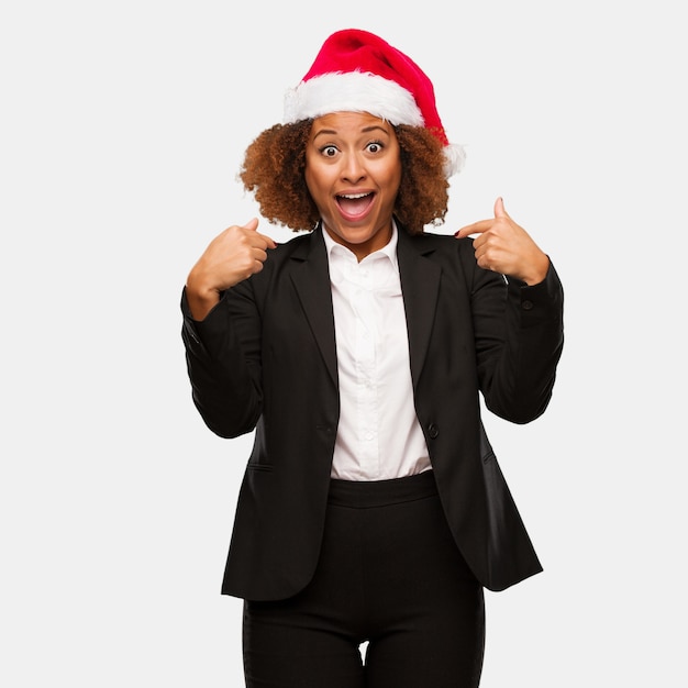 Young business black woman wearing a chirstmas santa hat surprised, feels successful and prosperous