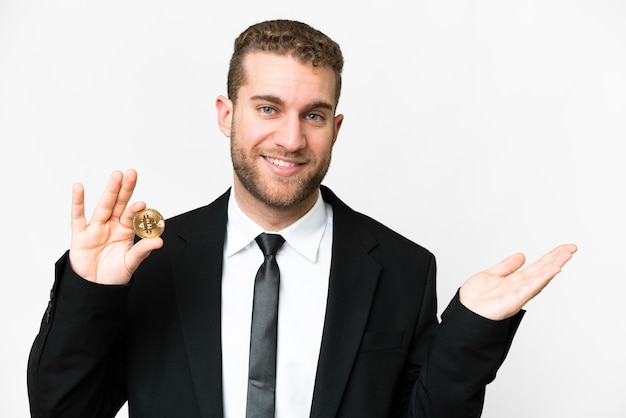 Foto giovane uomo biondo di affari che tiene un bitcoin sopra fondo bianco isolato che estende le mani al lato per invitare a venire