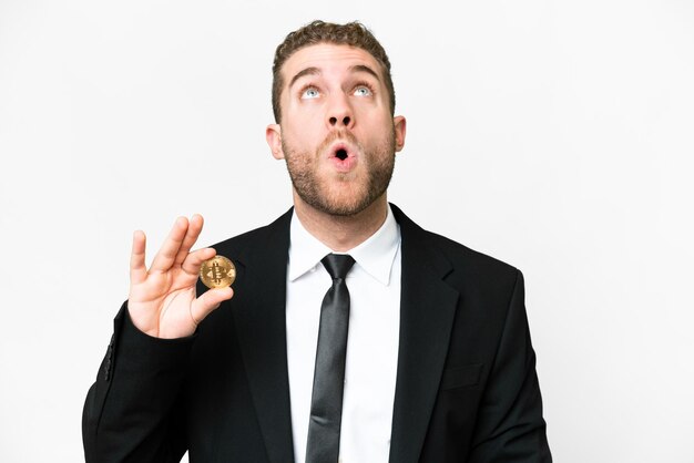 Young business blonde man holding a Bitcoin over isolated white background looking up and with surprised expression