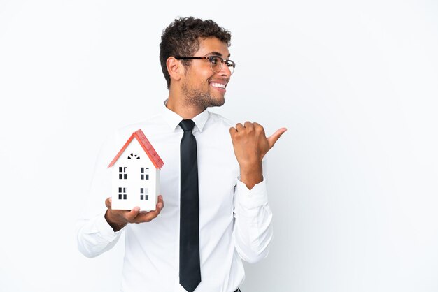 Young business Brazilian man holding a house toy isolated on white background pointing to the side to present a product