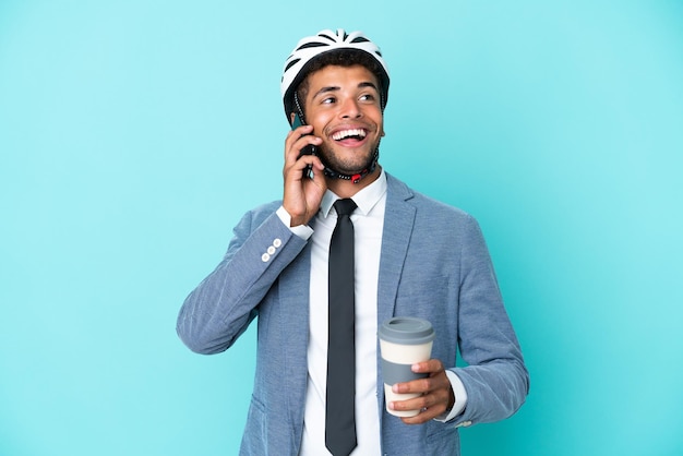 Young business Brazilian man with bike helmet isolated on blue background holding coffee to take away and a mobile