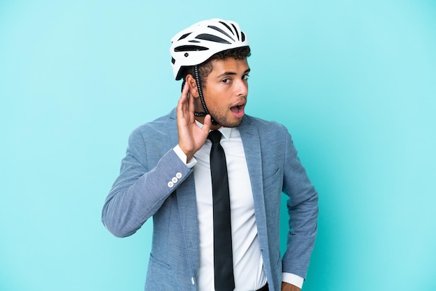 Молодой деловой бразильский мужчина с велосипедным шлемом изолирован на синем фоне, слушая что-то, положив руку на ухо