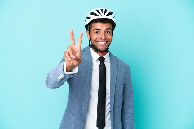 Giovane uomo brasiliano d'affari con casco da bici isolato su sfondo blu sorridente e che mostra il segno della vittoria