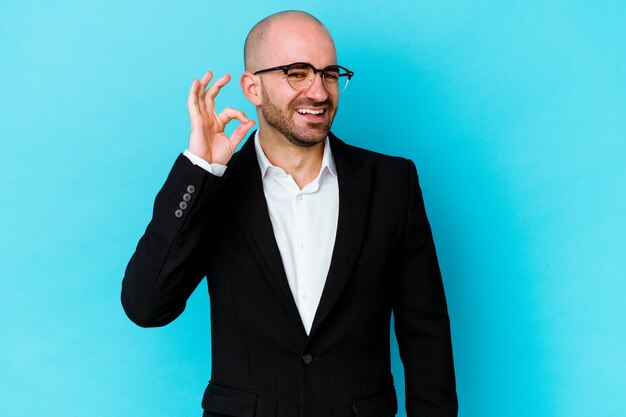 Young business caucasian bald man isolated on blue wall winks an eye and holds an okay gesture with hand