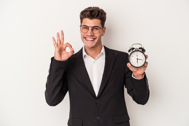 Young business caucasian man holding alarm clock isolated on white background cheerful and confident showing ok gesture.