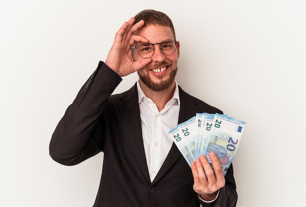 Young business caucasian man holding banknotes isolated on white background excited keeping ok gesture on eye.