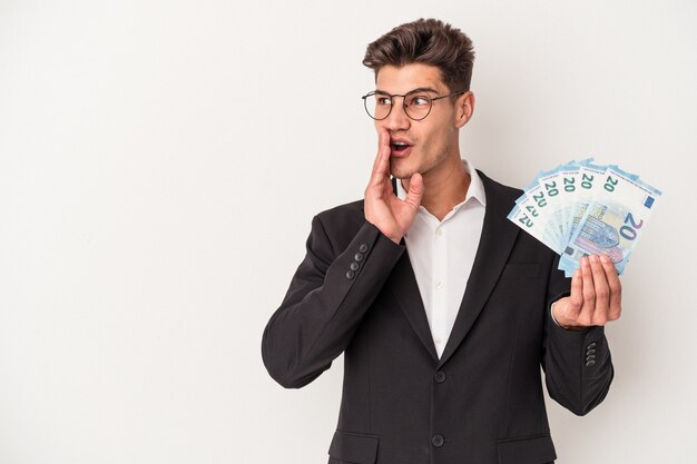 Young business caucasian man holding banknotes isolated on\
white background is saying a secret hot braking news and looking\
aside