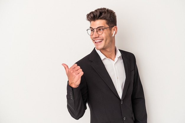 Young business caucasian man wearing headphones isolated on white background points with thumb finger away, laughing and carefree.