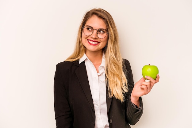 Young business caucasian woman holding an apple isolated on white background looks aside smiling cheerful and pleasant