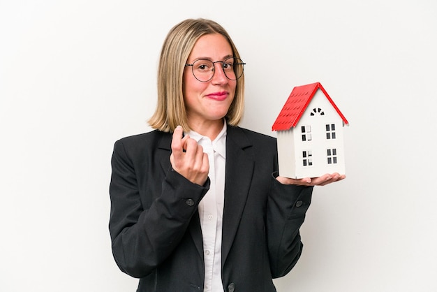 Young business caucasian woman holding a toy house isolated on white background pointing with finger at you as if inviting come closer