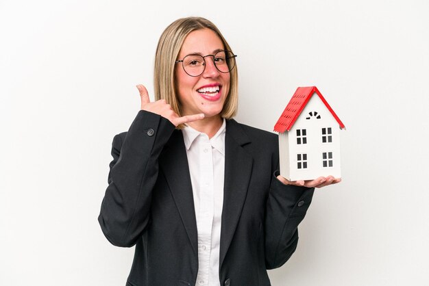 Young business caucasian woman holding a toy house isolated on white background showing a mobile phone call gesture with fingers