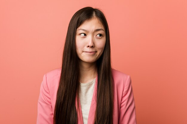 Young business chinese woman wearing pink suit confused, feels doubtful and unsure.