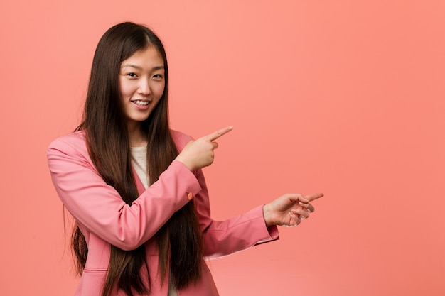 Young business chinese woman wearing pink suit excited pointing with forefingers away.