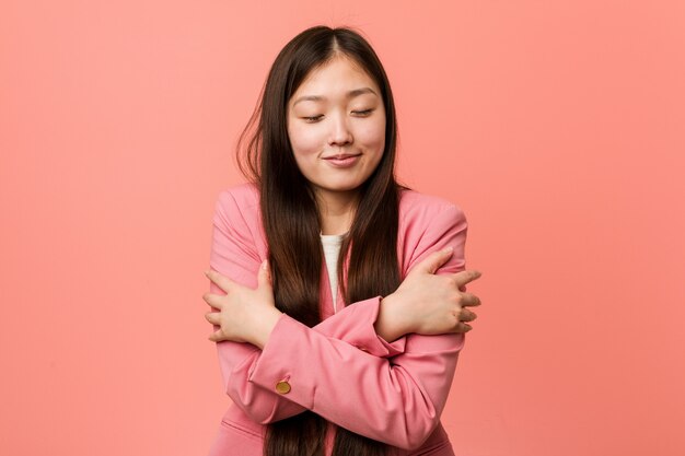 Young business chinese woman wearing pink suit hugs himself, smiling carefree and happy.