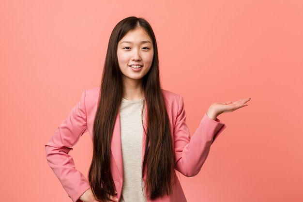 Young business chinese woman wearing pink suit showing a copy space on a palm and holding another hand on waist.