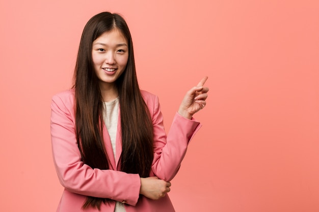陽気に離れて人差し指で指している笑顔ピンクのスーツを着ている若いビジネス中国女性。