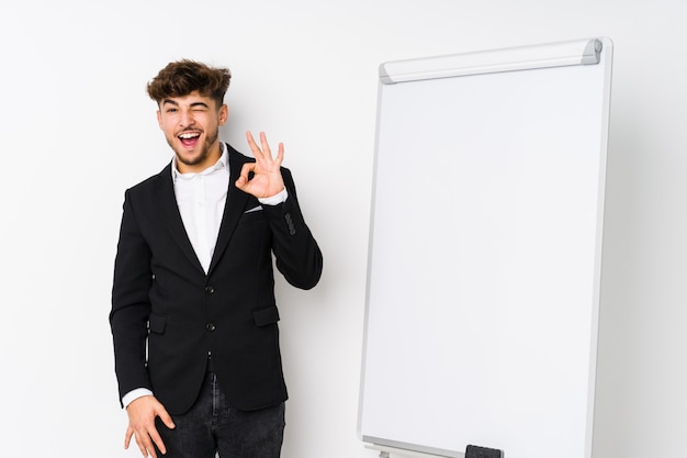 Young business coaching arabian man winks an eye and holds an okay gesture with hand.