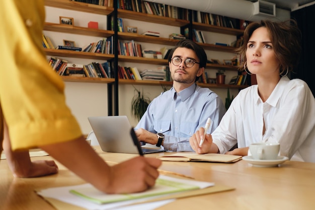 Young business colleagues sitting at the desk with laptop thoughtfully looking at boss while working with papers in modern office