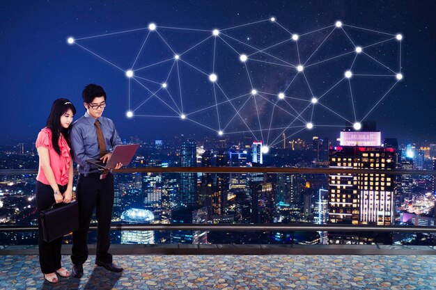 Young business couple using a laptop while working together on the office rooftop with connection network above modern city at night time