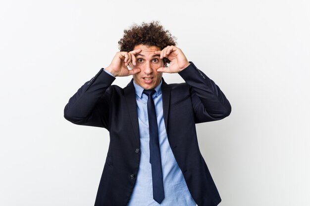 Young business curly man against white wall keeping eyes opened to find a success opportunity.