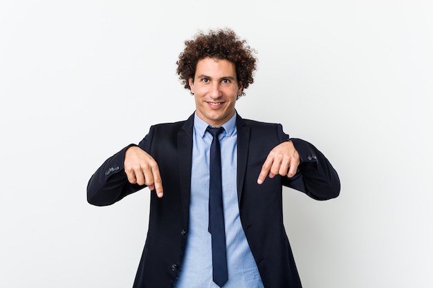 Young business curly man against white wall points down with fingers, positive feeling.