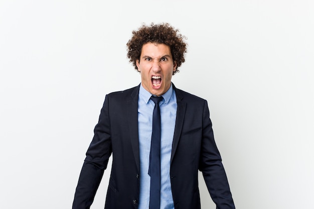 Young business curly man against white wall screaming very angry and aggressive.