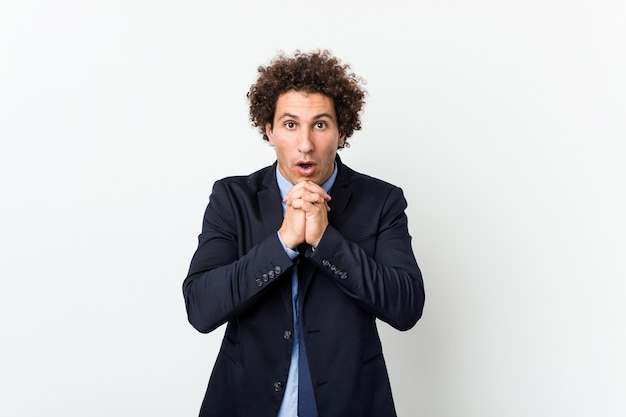 Young business curly man on white background praying for luck, amazed and opening mouth looking to front.