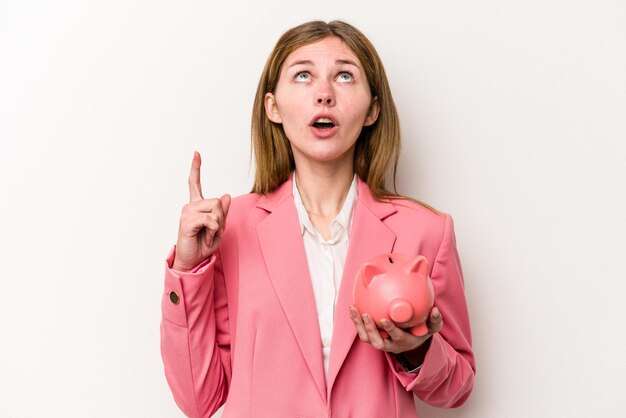 Young business English woman holding a piggybank isolated on white background pointing upside with opened mouth.