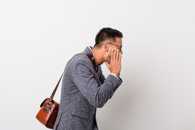 Young business filipino man against a white wall shouts loud, keeps eyes opened and hands tense.