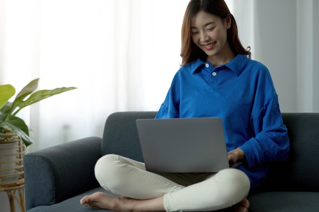 젊은 비즈니스 프리랜서 아시아 여성이 집 거실에서 휴식을 취할 때 소파에 누워 소셜 미디어를 확인하면서 노트북 작업을 하고 있습니다.