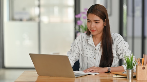 ノートパソコンに入力して木製の作業机に座ってストライプのシャツの若いビジネスの女の子