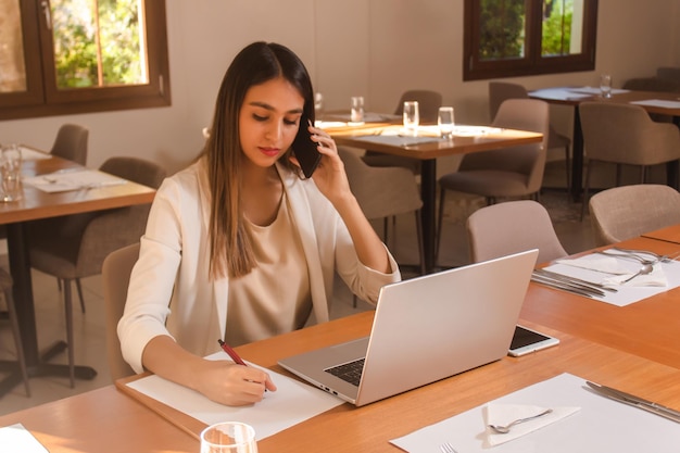 Young business girl talking on the phone while taking notes in a restaurant