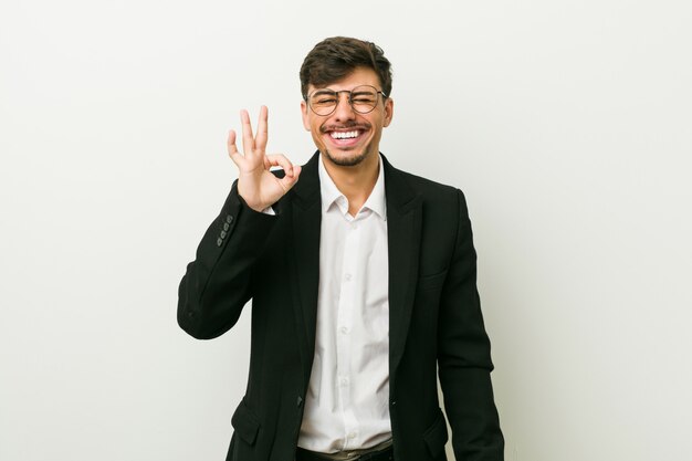 Young business hispanic man winks an eye and holds an okay gesture with hand.