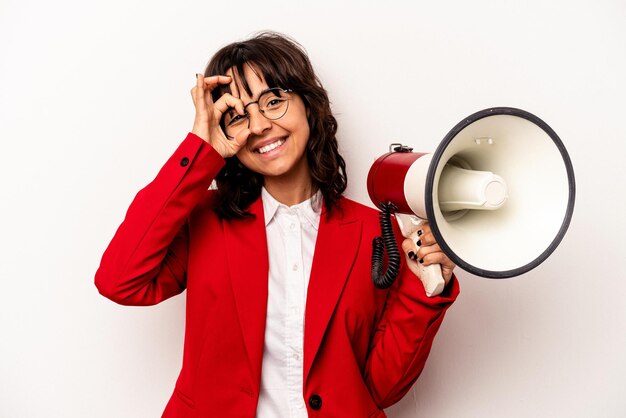 Young business hispanic woman holding an megaphone isolated on white background excited keeping ok gesture on eye